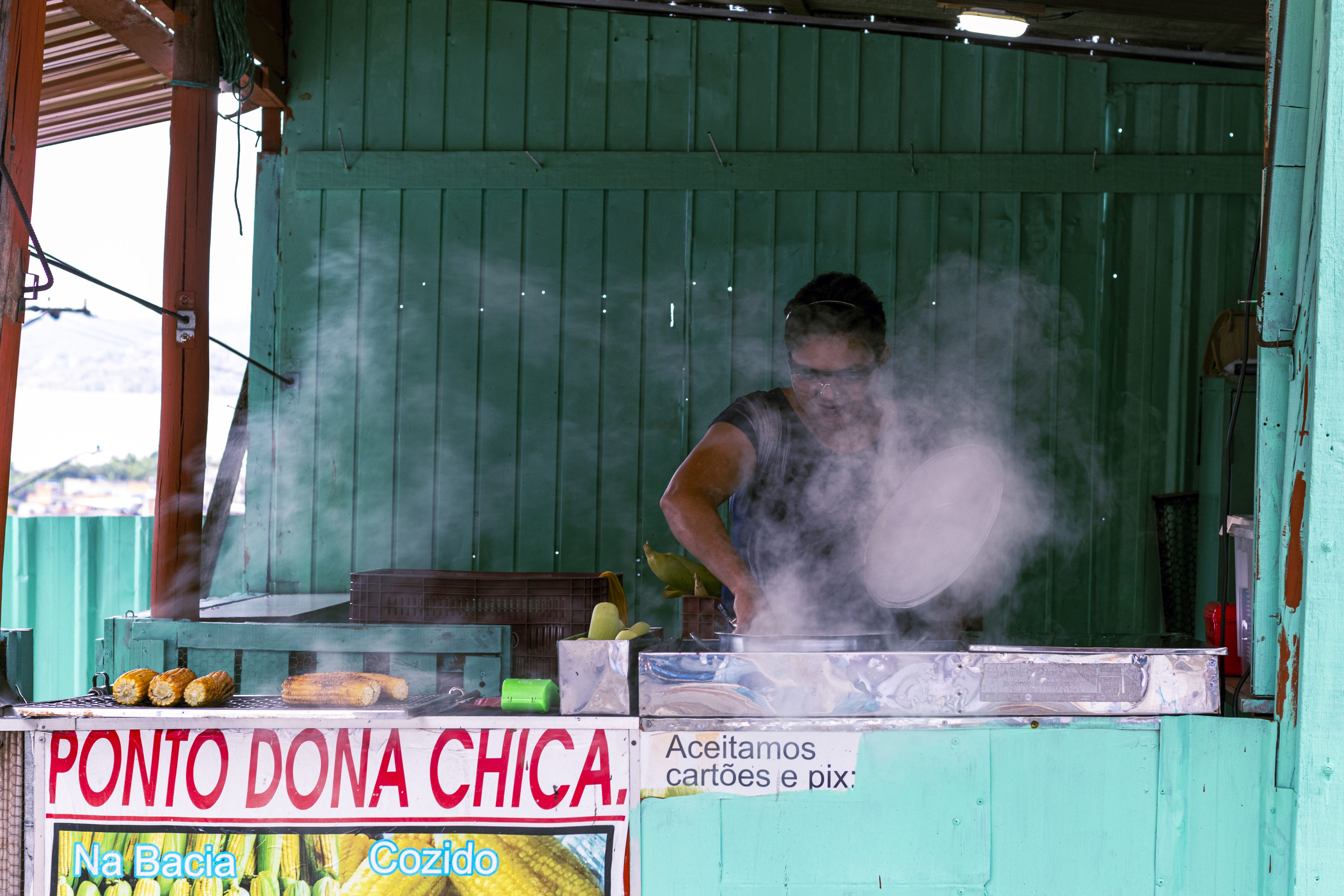A vendedora de milho está exposta a um calor intenso por conta do fogo e cinzas do carvão e, por conta disso, usa um boné e um óculos de proteção.